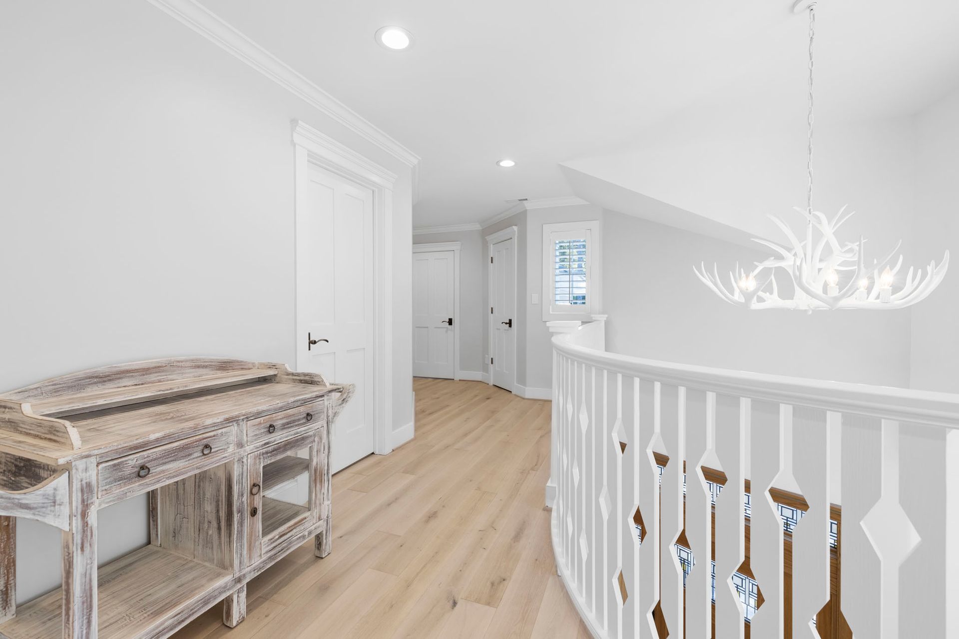 Bright hallway with white walls, light wood floors, a rustic console table, and an upstairs railing with a chandelier.