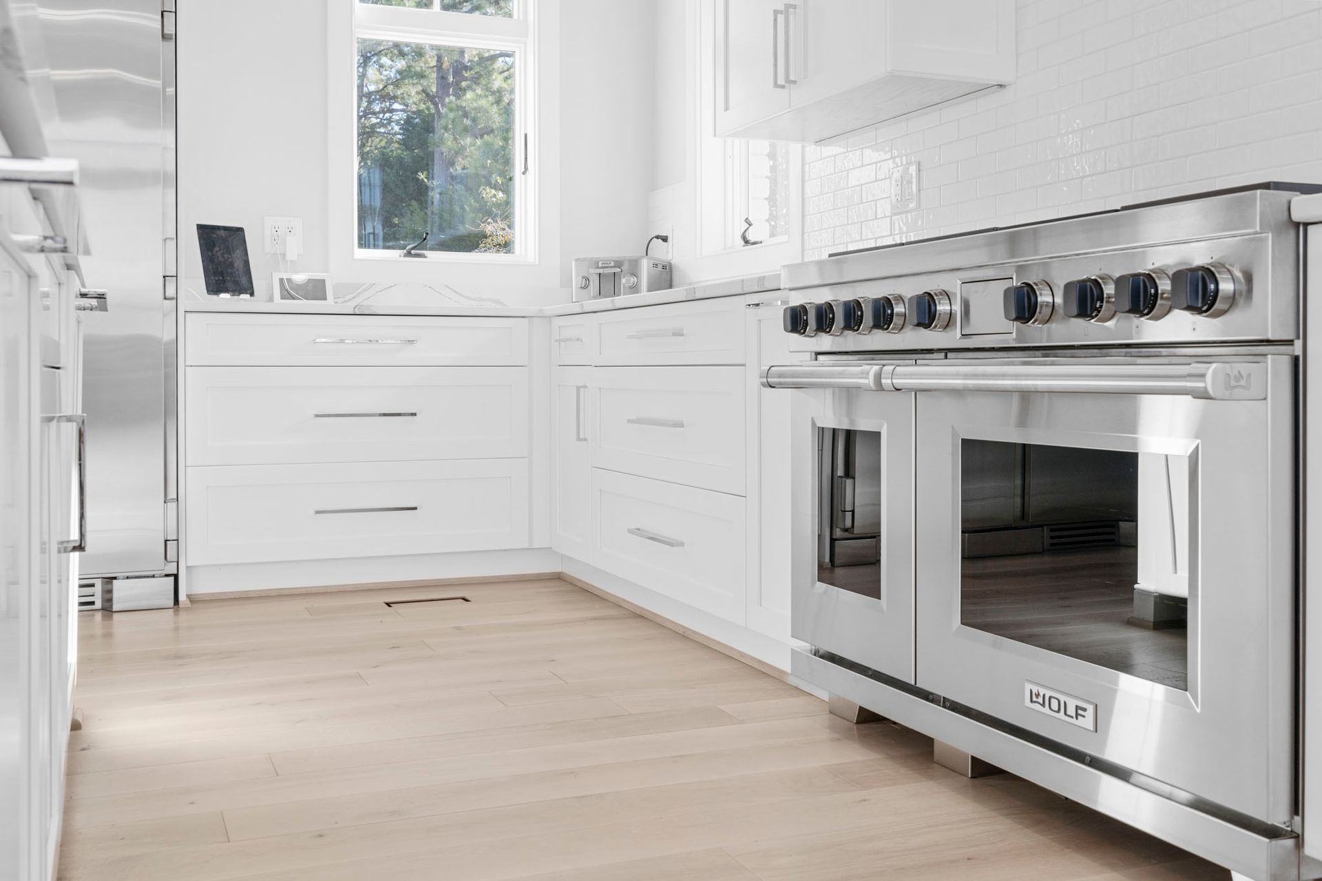Bright modern white kitchen with stainless steel oven, light wood floor, and a window over the sink