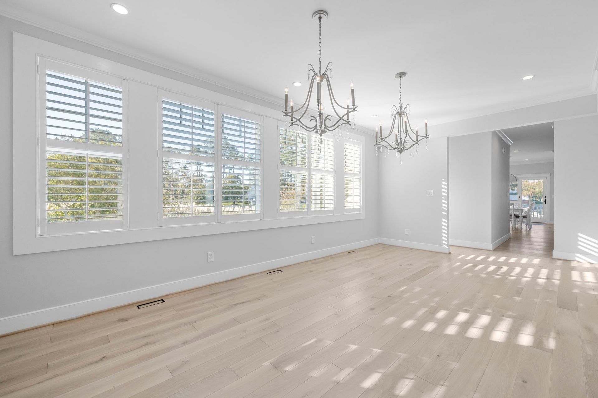 Bright empty room with hardwood floors, white shutters, and two chandeliers