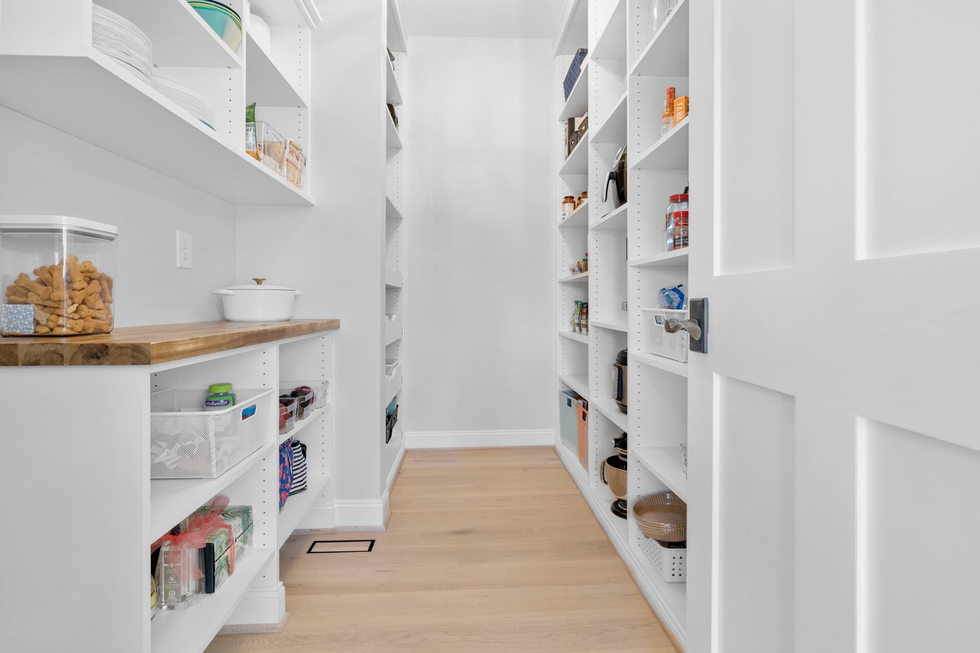 Bright white walk-in pantry with wooden shelves, woven baskets, and a light wood floor