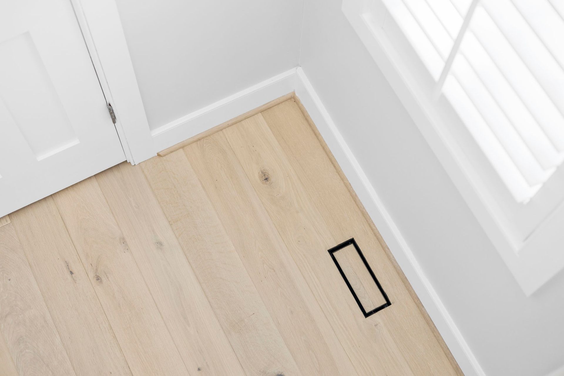 Light wood floor by a white wall and door, with a black rectangular floor vent or cover.