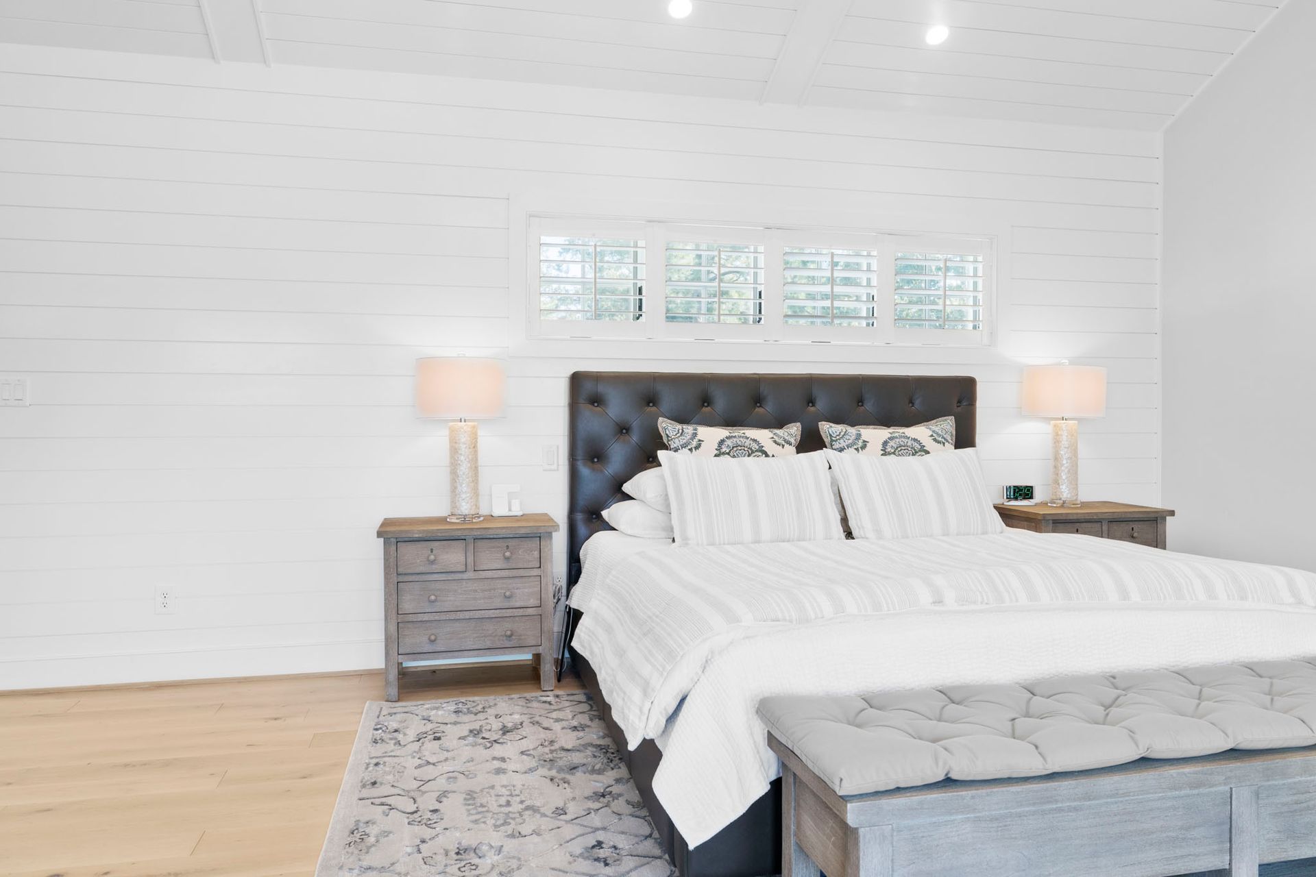 Bright white bedroom with a tufted dark headboard, neatly made bed, nightstands, and lamps