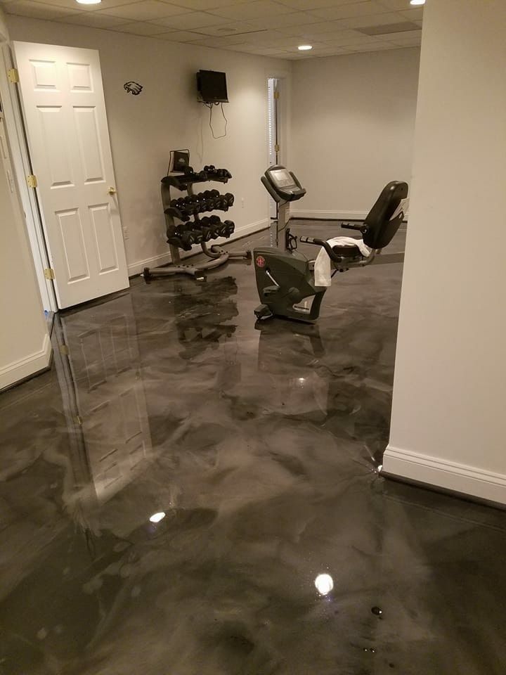 Basement gym with a stationary bike, dumbbells, mirror, and glossy gray floor.