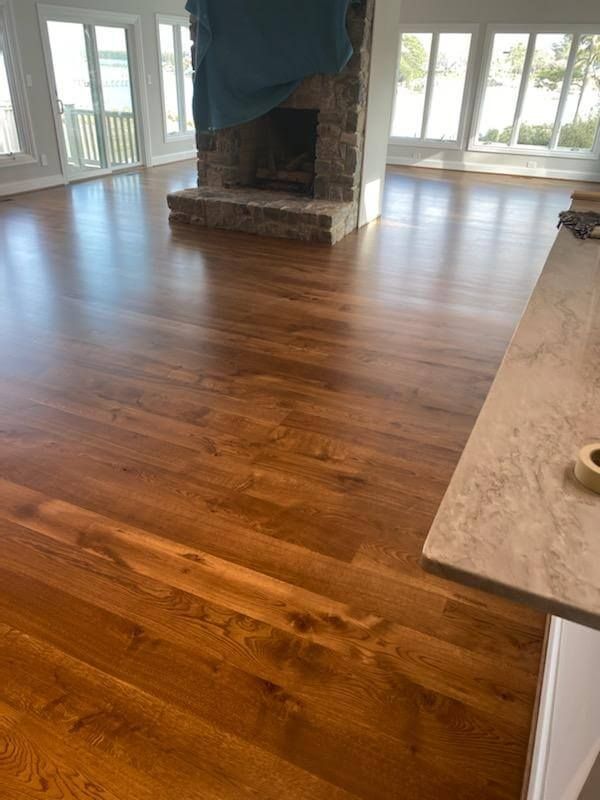 Empty living room with hardwood floors, a stone fireplace, and large windows letting in daylight