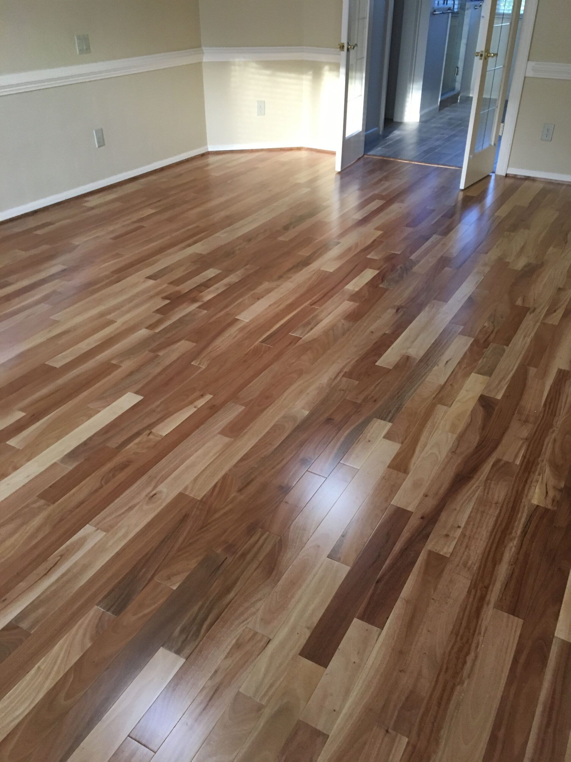 Empty room with shiny wood plank flooring and beige walls, lit by daylight from a hallway.