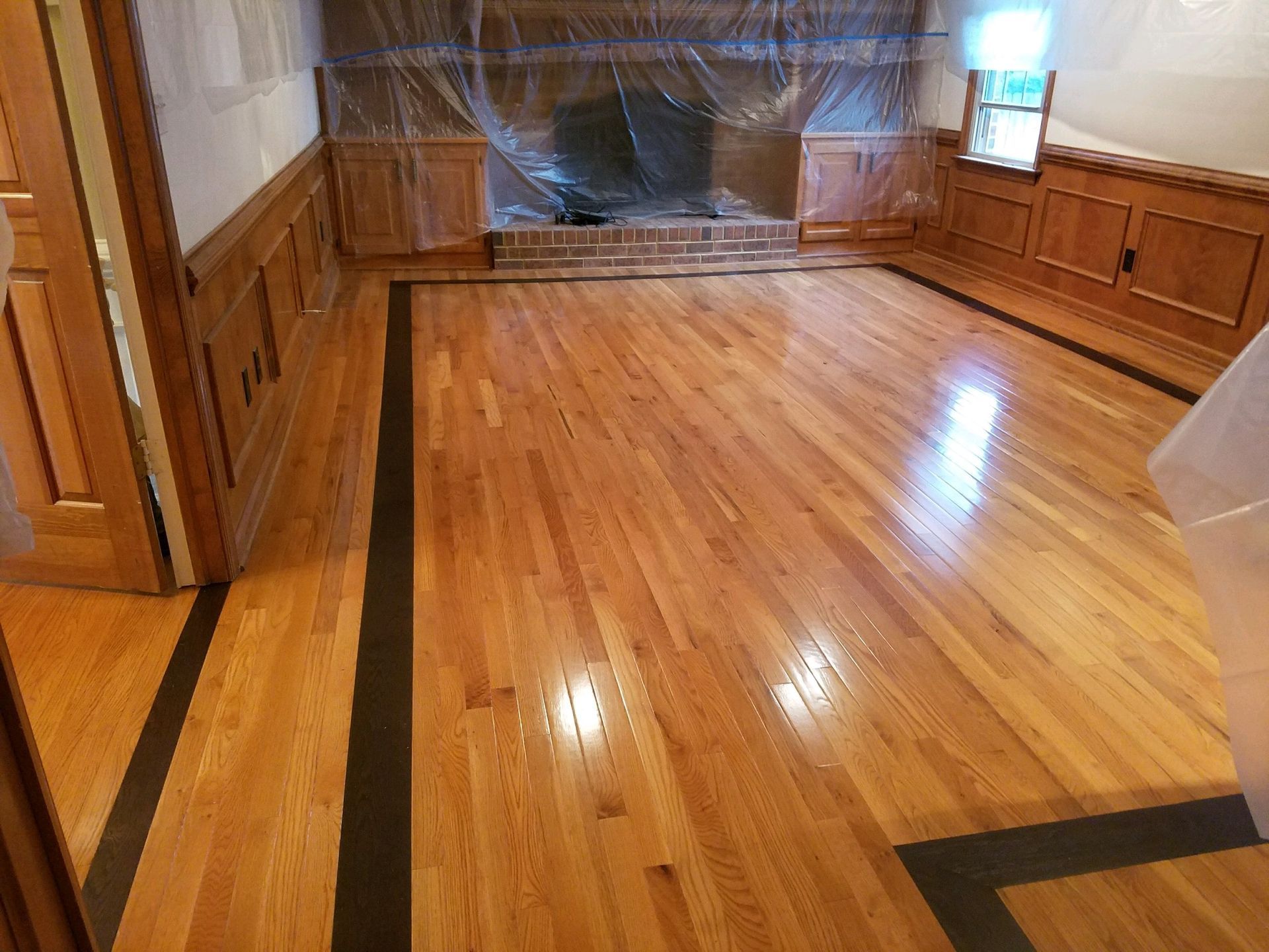Empty wood-paneled room with polished hardwood floors and a windowed back wall covered in plastic.