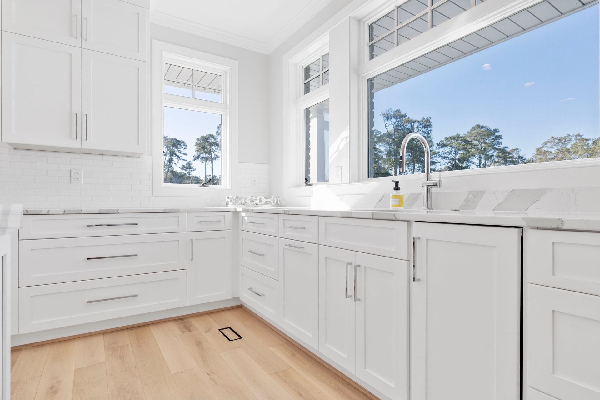 Bright white kitchen with cabinets, wood floor, and large windows overlooking trees and blue sky