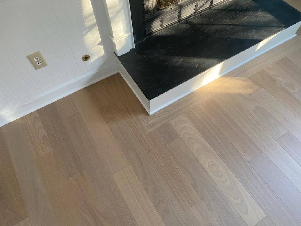 Empty room with light wood flooring, white wall, and black tiled hearth by a window.