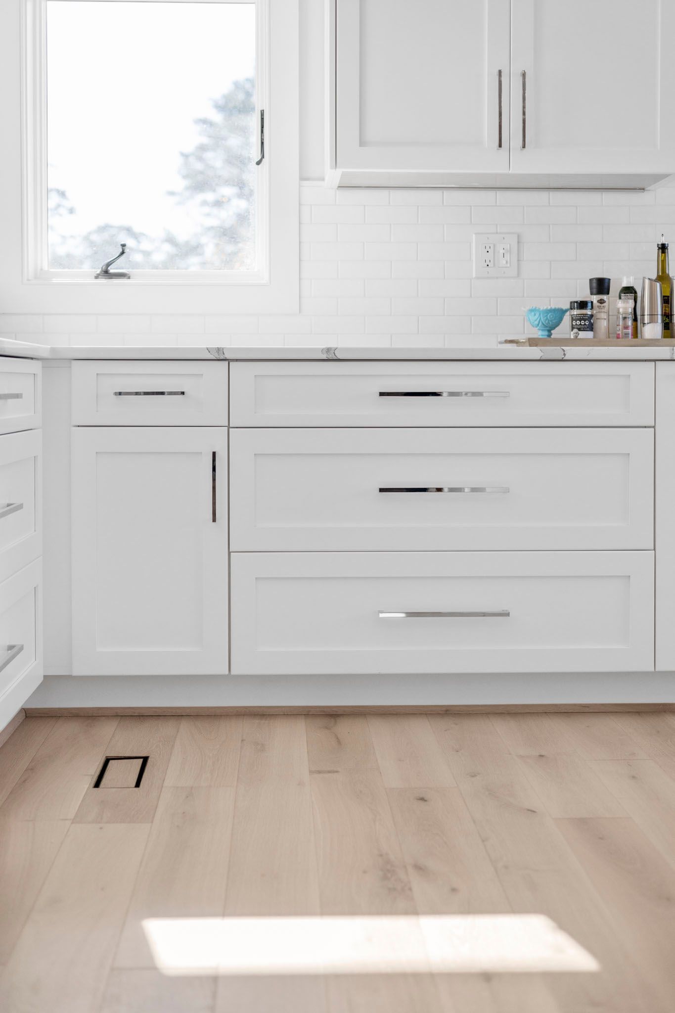 Bright white kitchen with cabinets, a window, and light wood floor, sunlit by a patch of light.