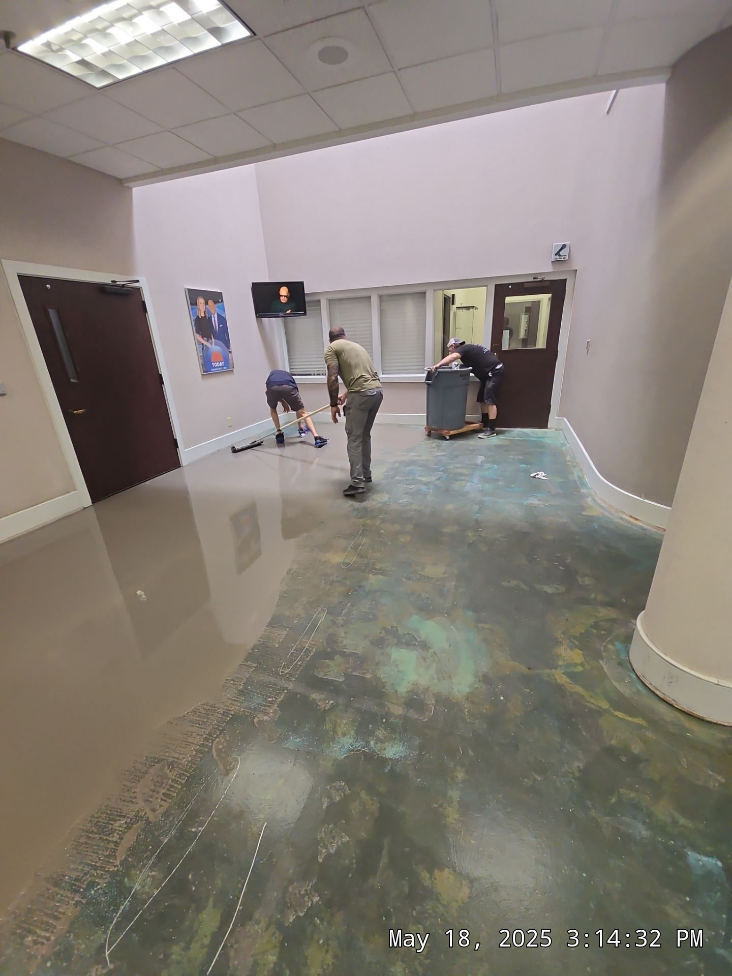 Spacious lobby with glossy floor; three people near a wall and doors, one bending over a bag.