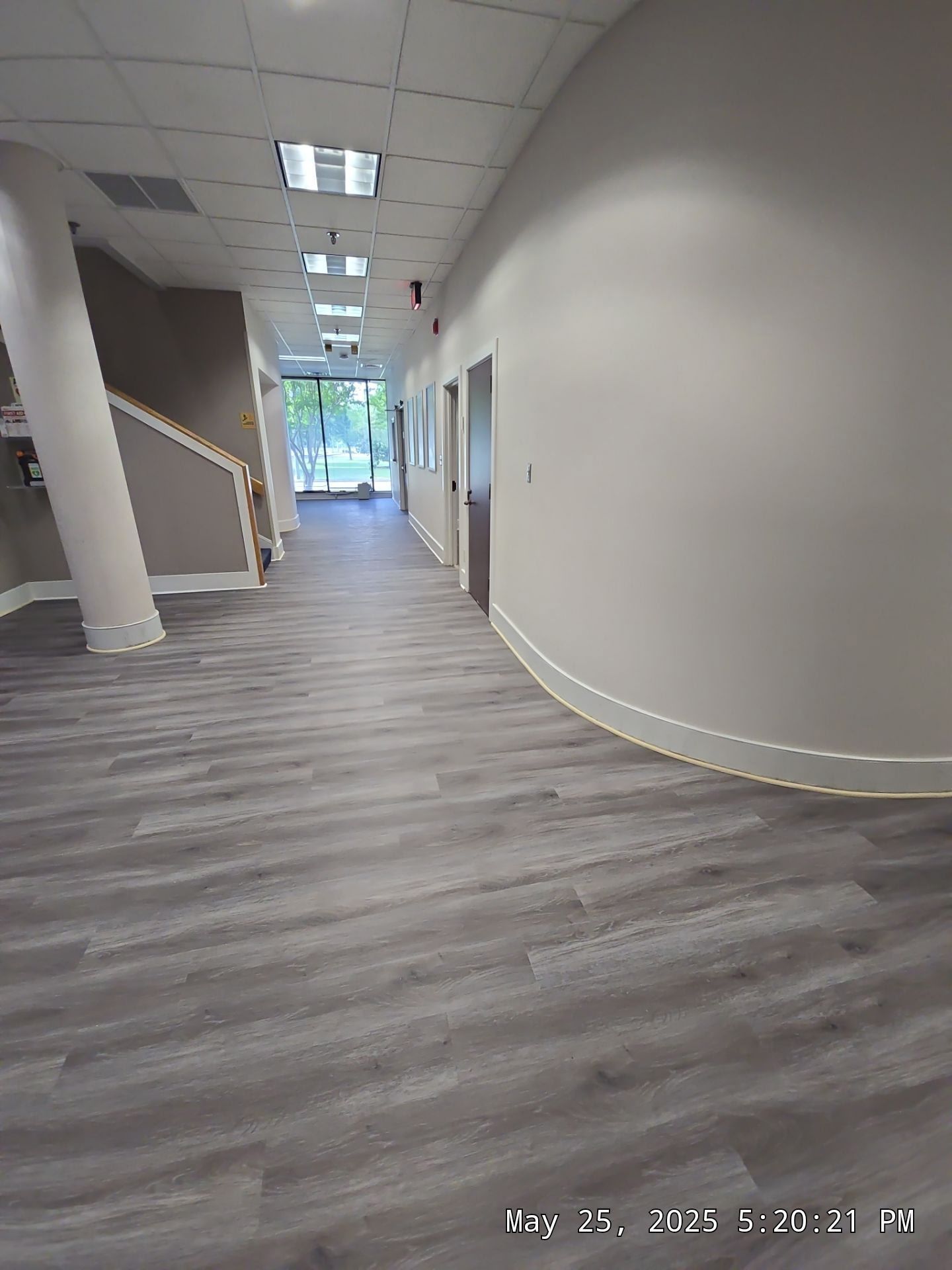 Empty office hallway with gray flooring, white walls, stairs on the left, and bright windows at the end.