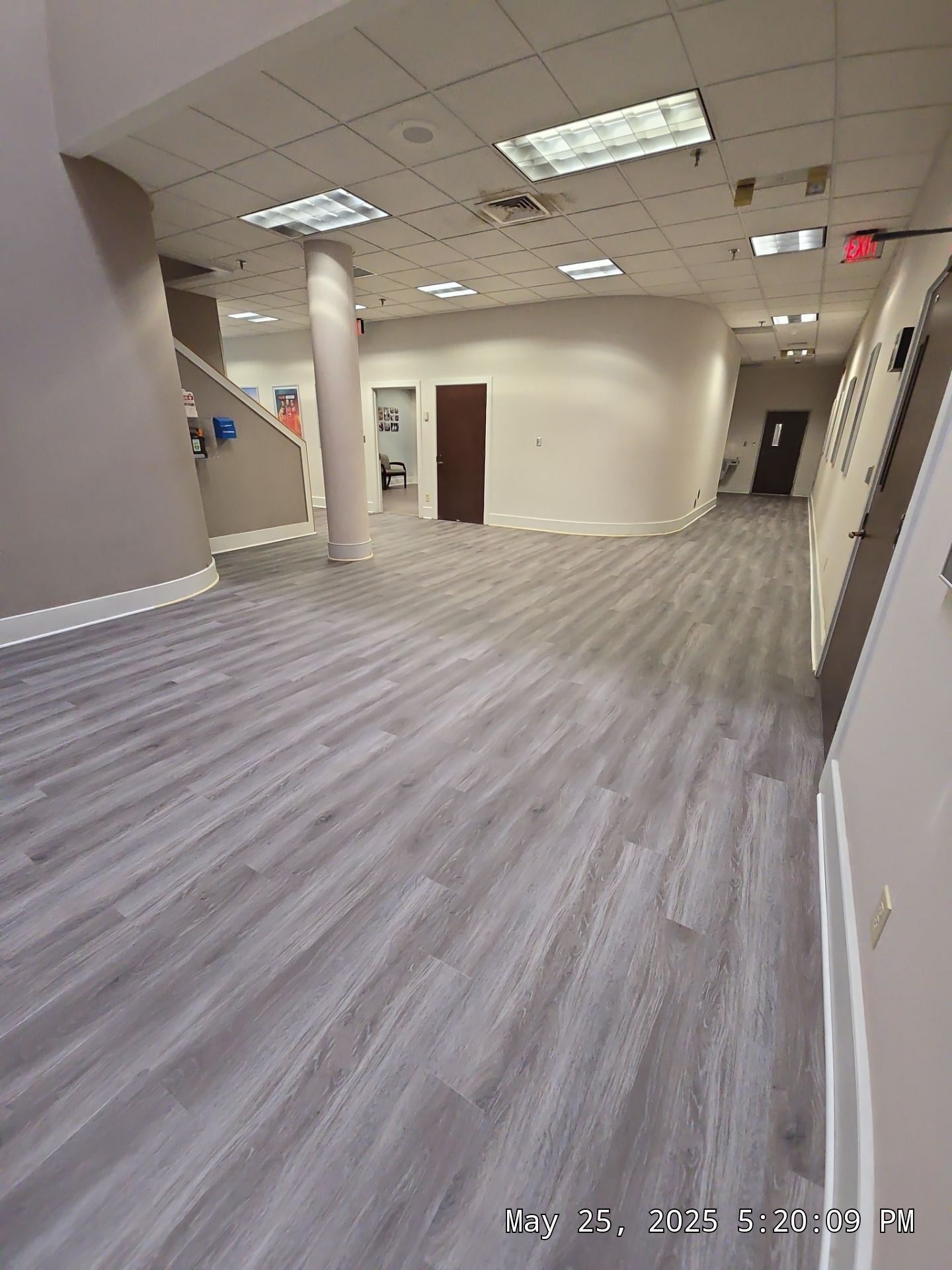 Empty office hallway with gray wood flooring, beige walls, white columns, and ceiling lights