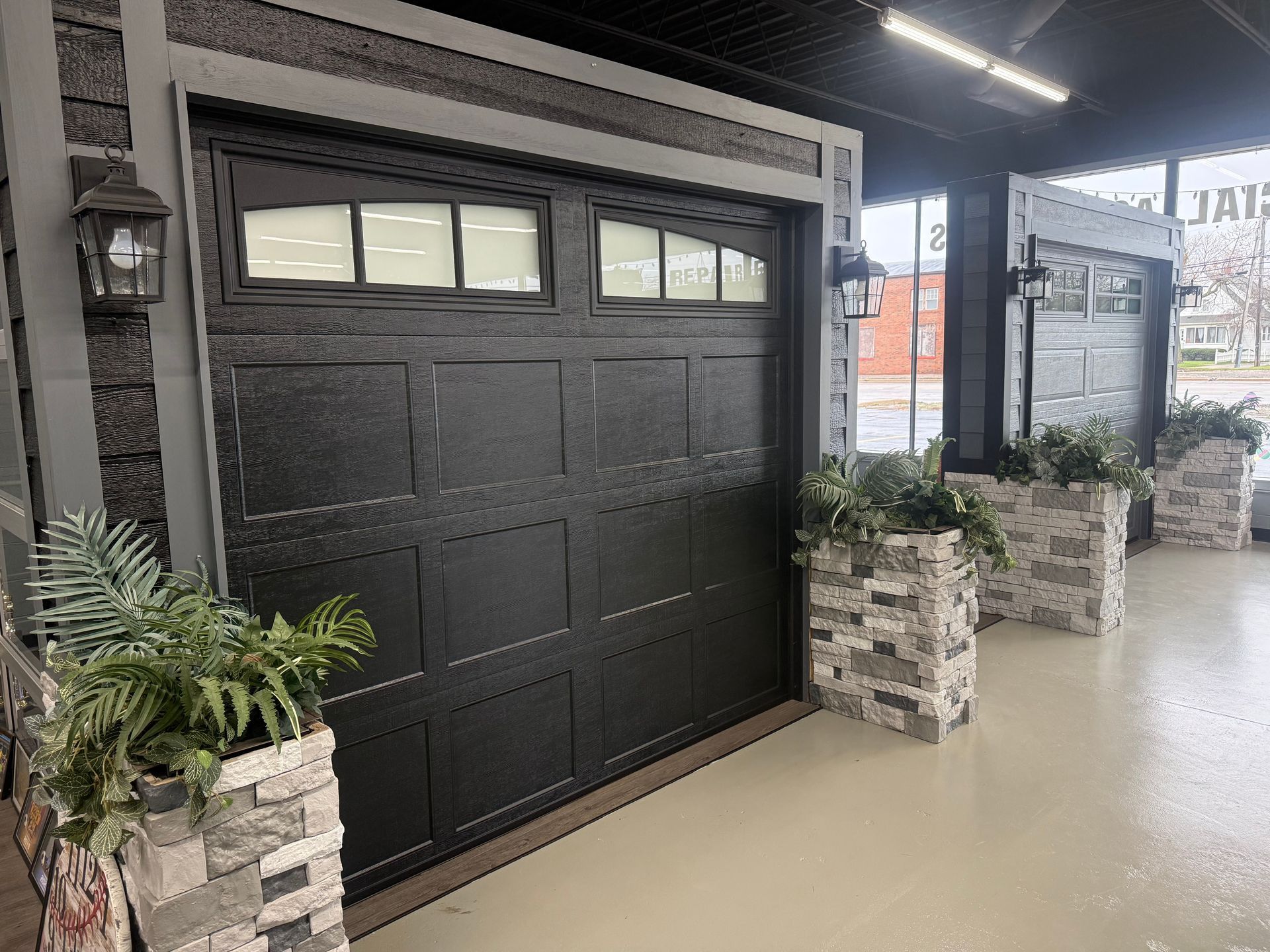 A showroom display featuring a black, paneled garage door with windows, flanked by stone planters filled with greenery.