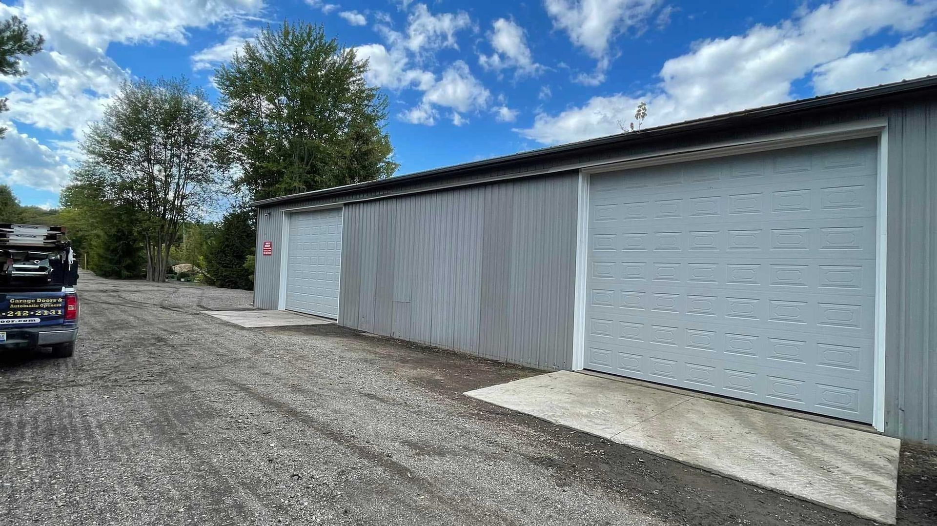 Gray storage units with roll-up doors, gravel driveway, and a partially visible blue sky.