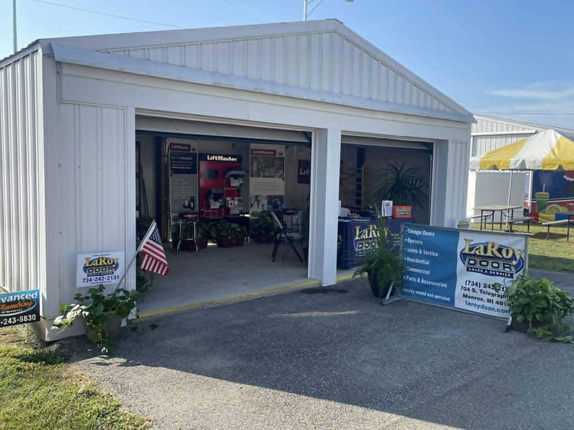 White two-bay garage with open doors at an outdoor event. Signage, plants, and an American flag are visible.
