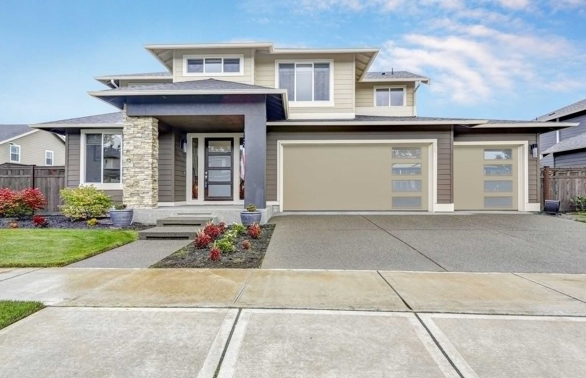 Modern two-story house with beige garage doors and trim, concrete driveway, and front yard landscaping under a blue sky.