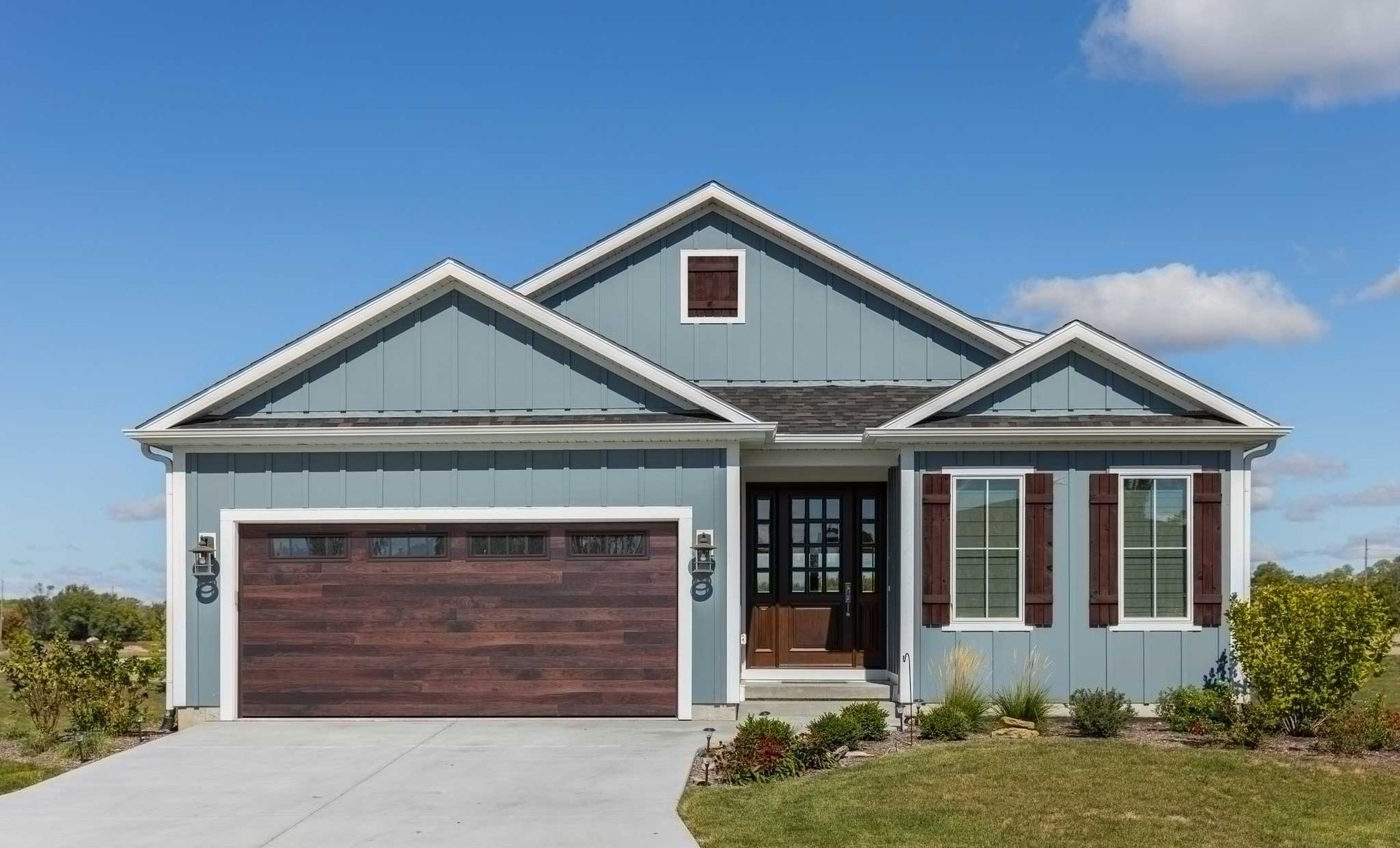 Blue-sided house with brown garage door, front door, and shutters; concrete driveway, green grass, blue sky.