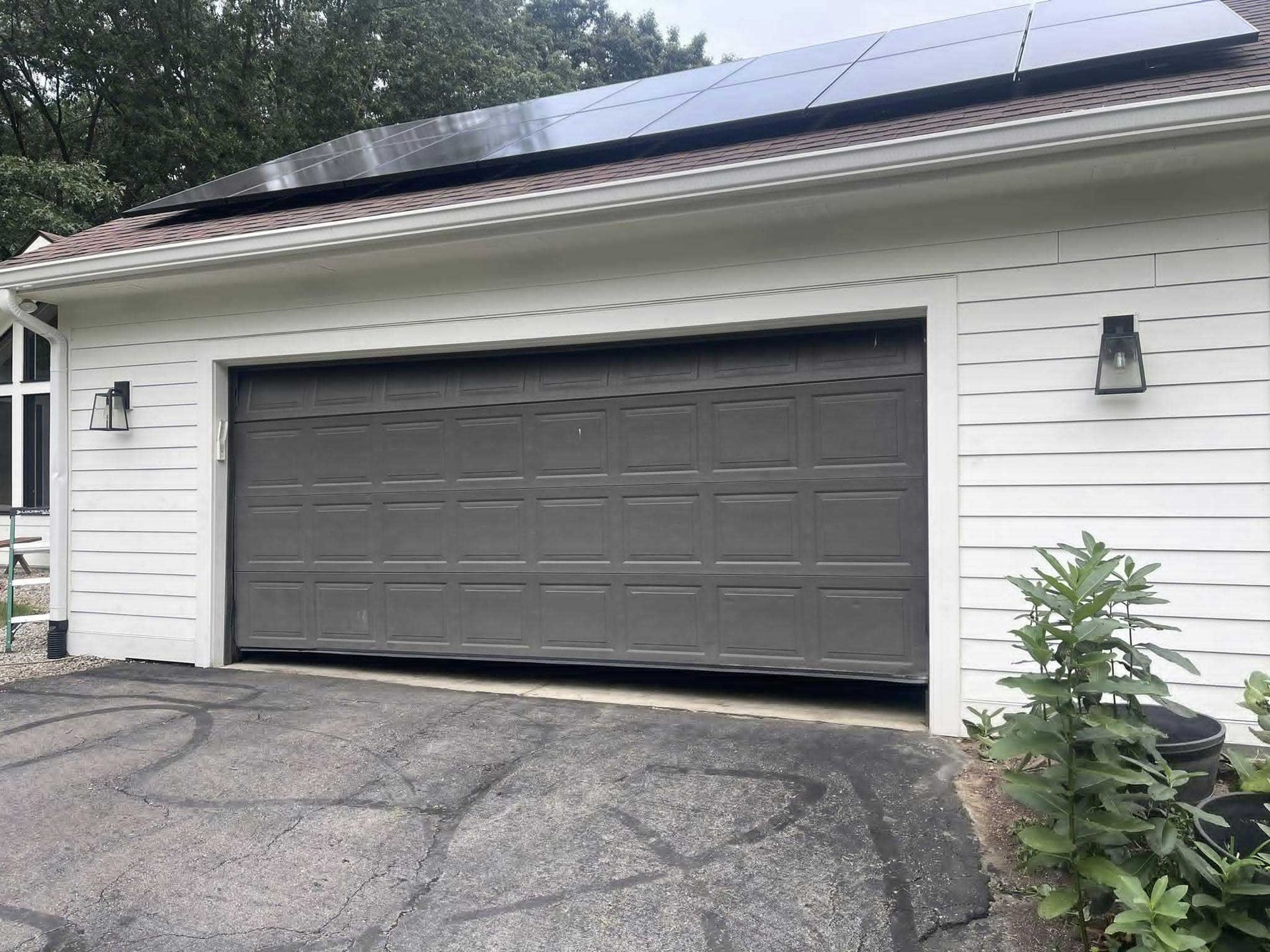 Gray garage door with solar panels on roof. White siding and a paved driveway.
