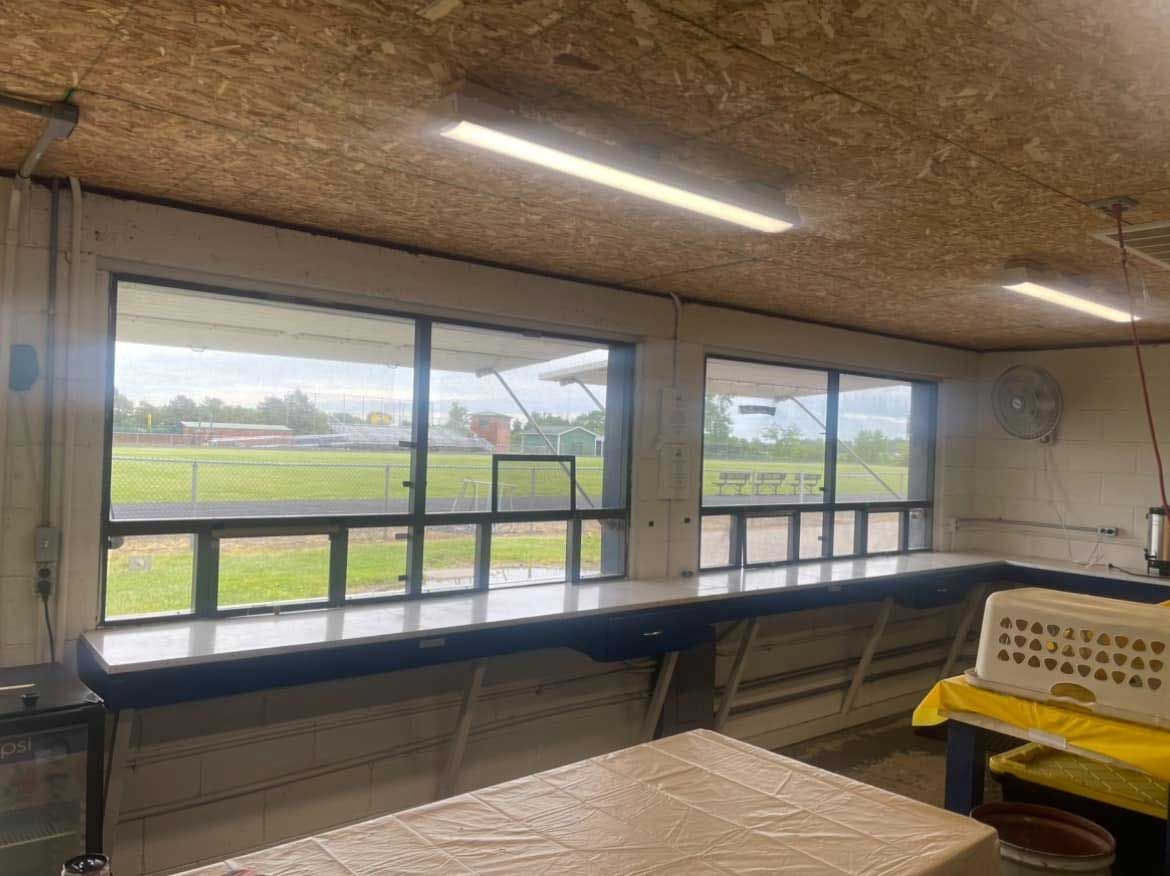 Interior of a concession stand, with large windows overlooking a green field. Long counter, fluorescent lights, and a table.