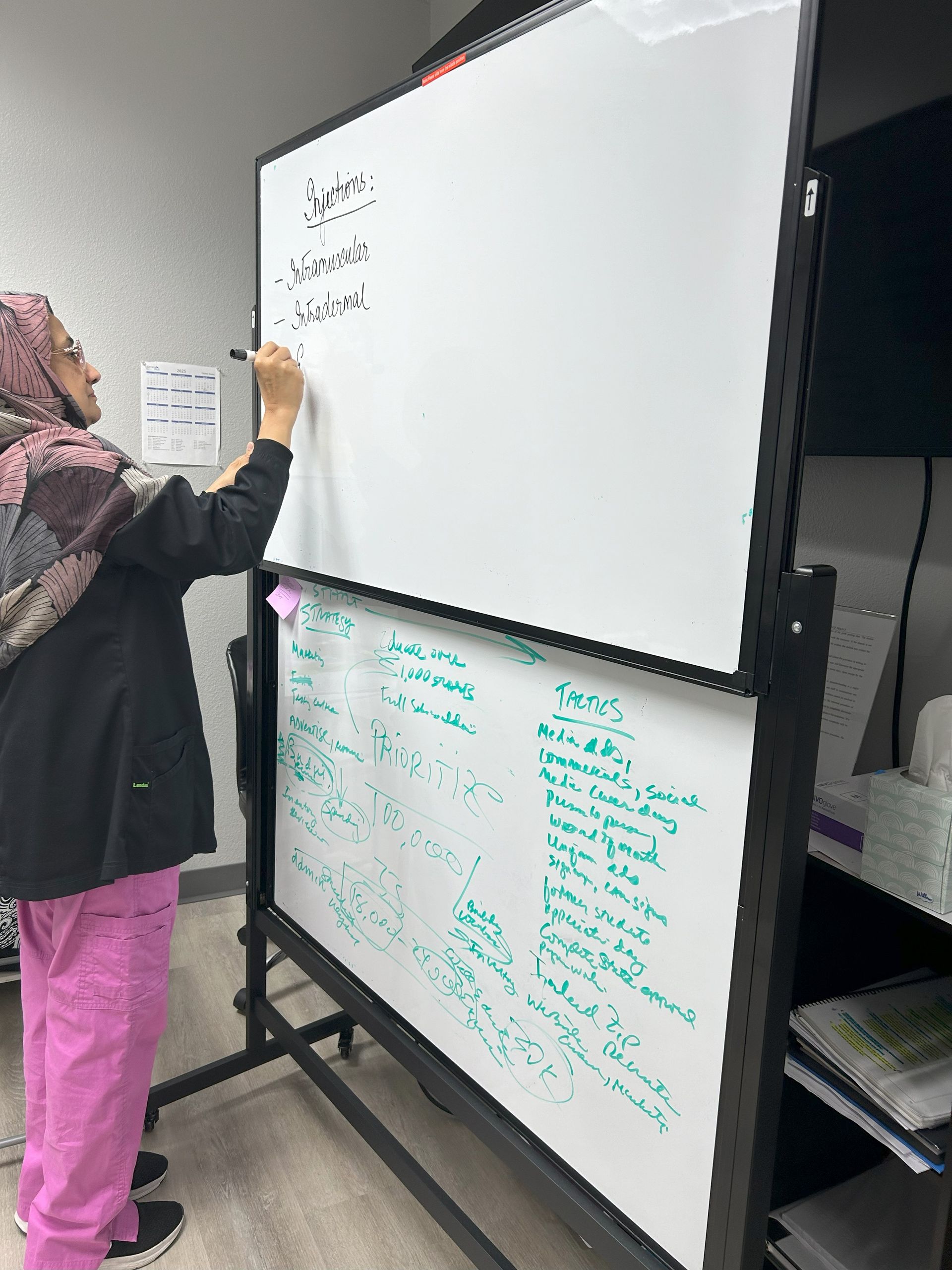 A woman in pink scrubs is writing on a whiteboard.