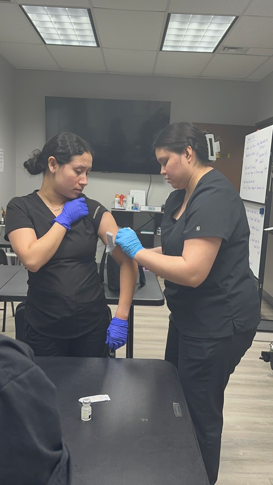 Two nurses are working on a patient's arm in a classroom.
