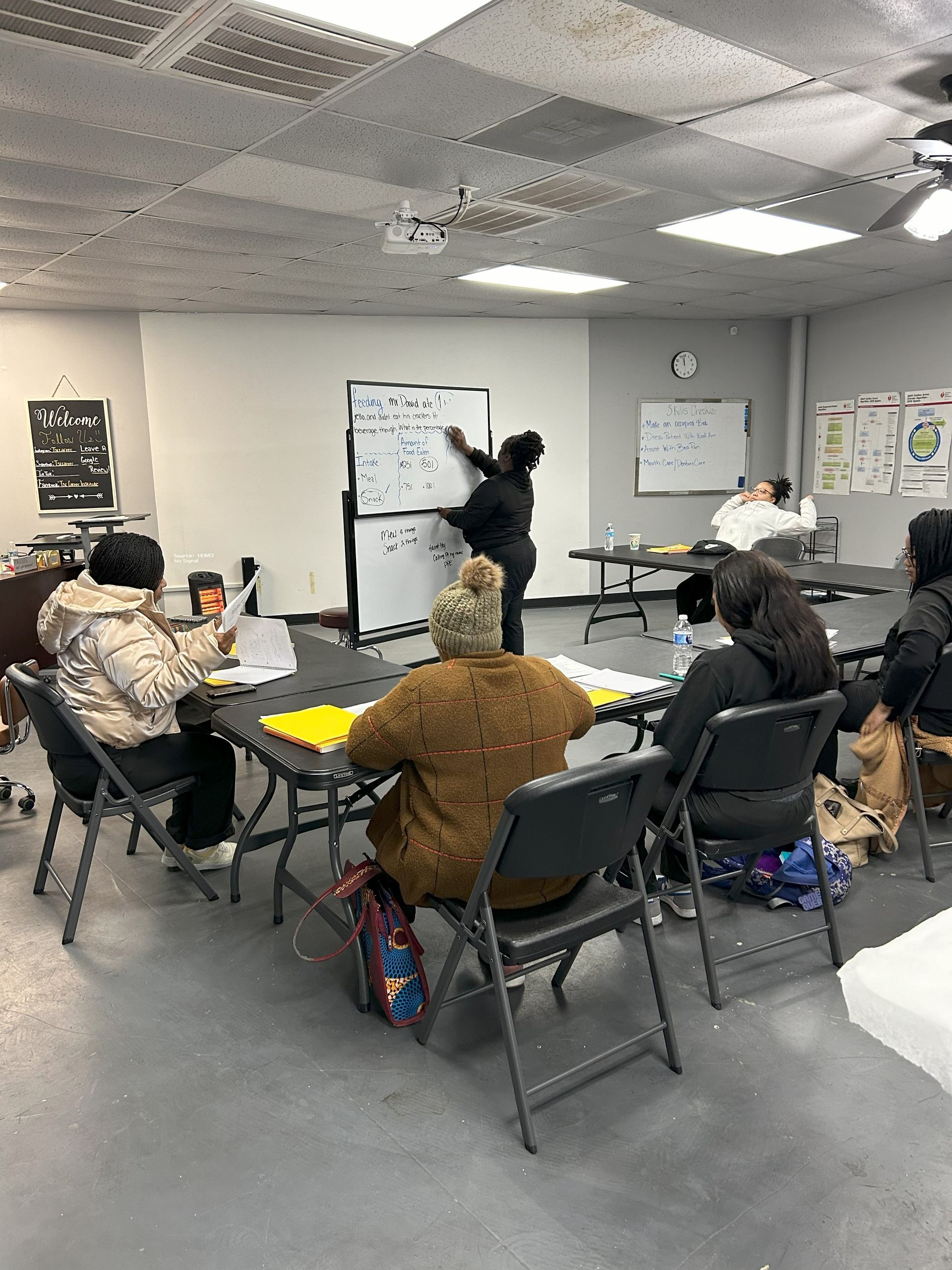 A group of people are sitting at tables in a room with a whiteboard.