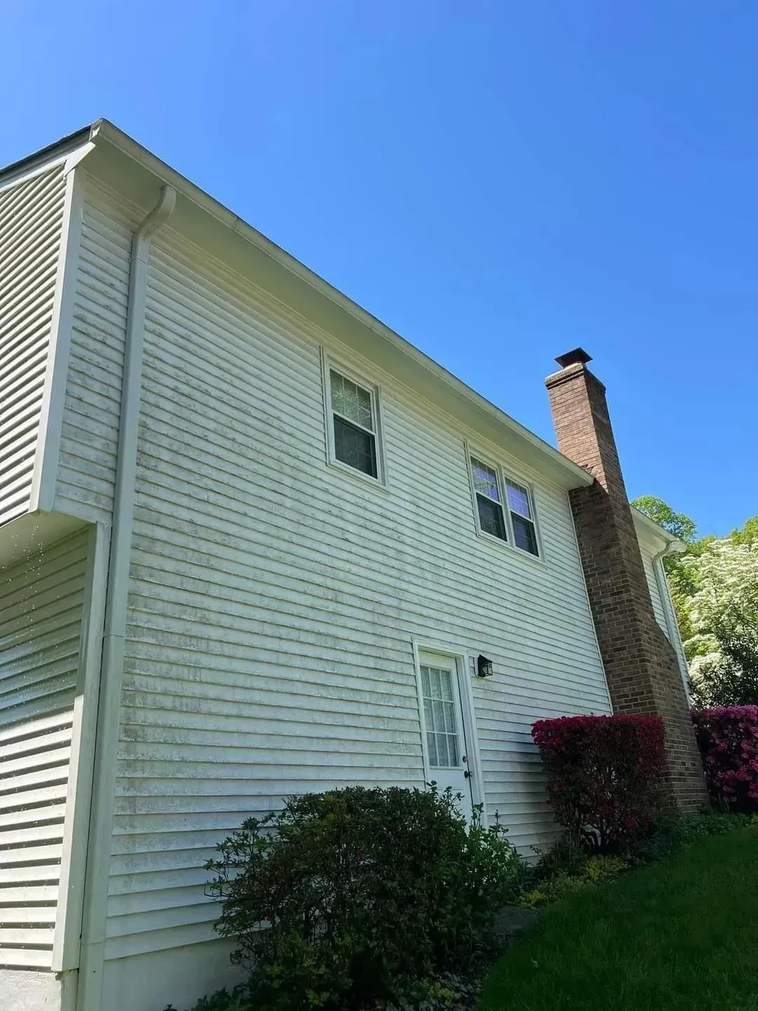 White-sided house with visible algae, brick chimney, windows, and door, under a blue sky.