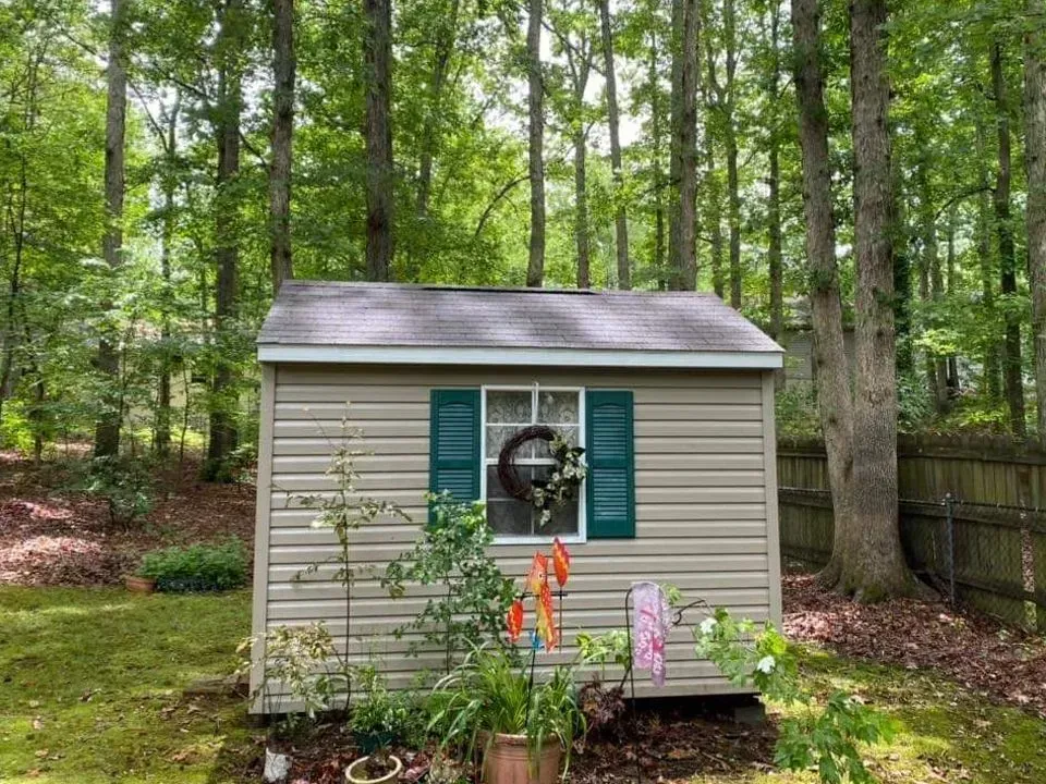 Tan shed with green shutters and a wreath in the window, surrounded by trees in a yard.