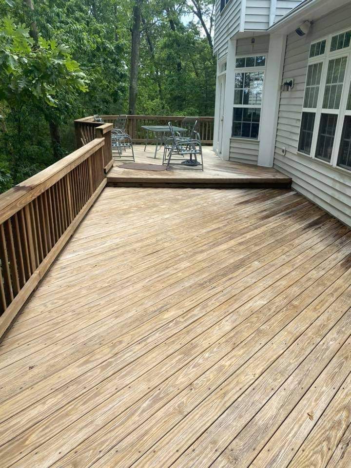 Wooden deck with railings, connected to a white house with windows. Forest in the background.