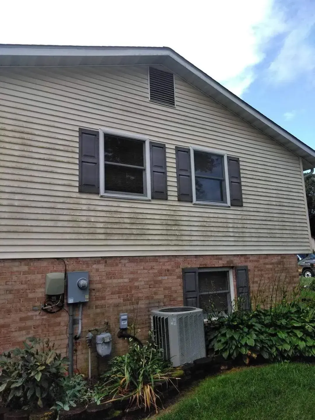 Side of a house with light tan siding, brick base, and brown shutters. There are two windows and an air conditioning unit.