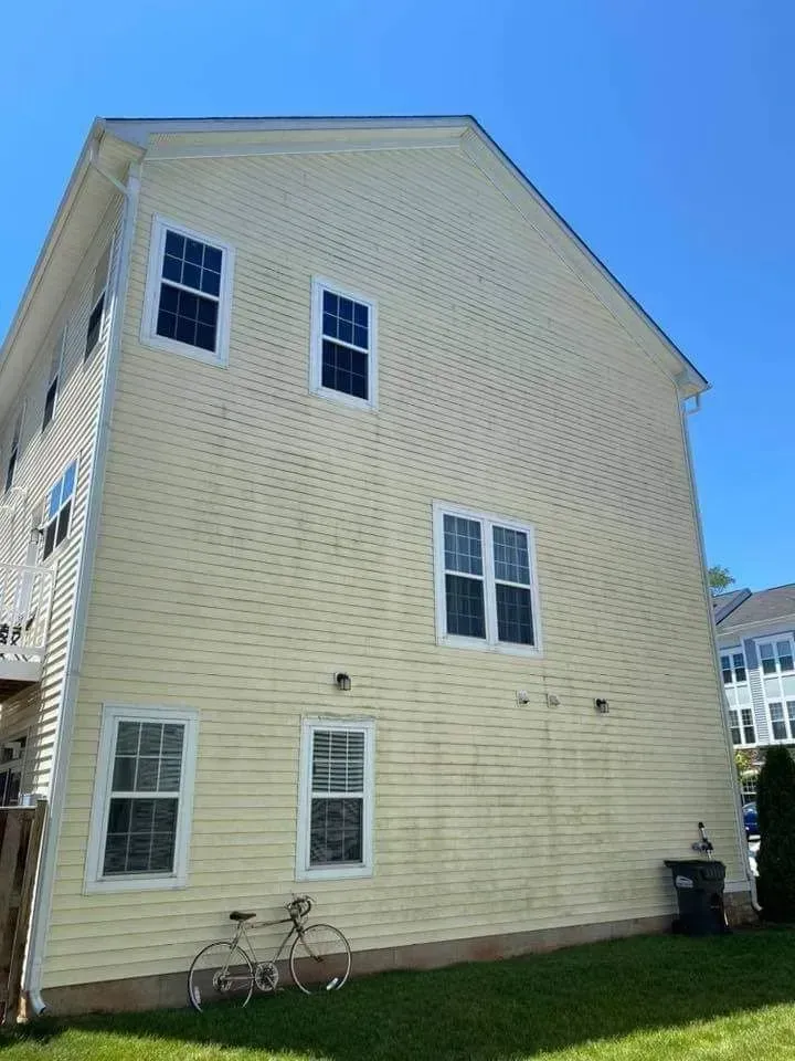Yellow-sided two-story house with white-framed windows, bicycle, and trash can on a sunny day.