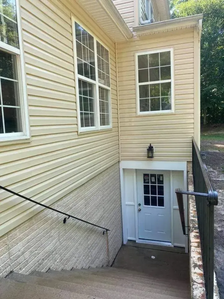 Exterior view of a building with a set of stairs leading to a white door. Tan siding and windows are visible.