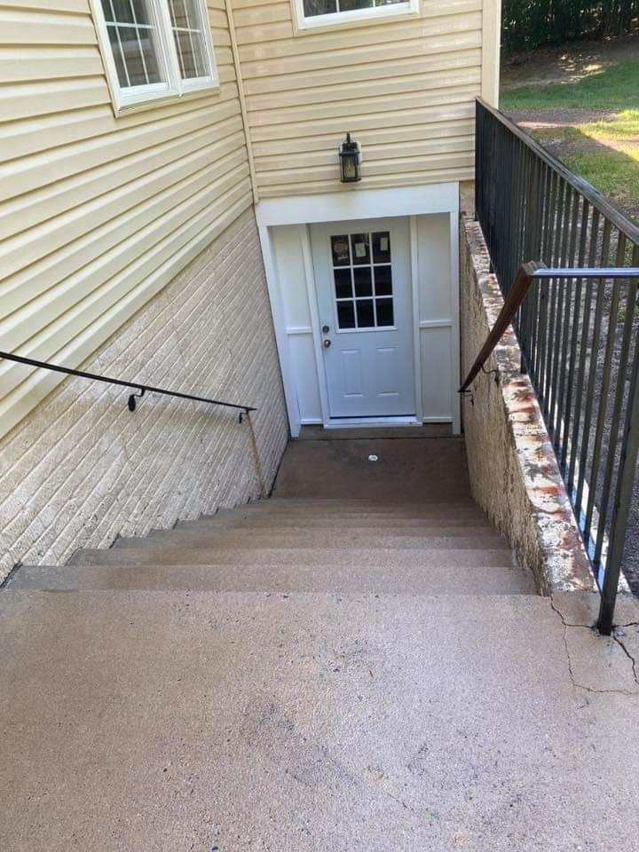 Concrete stairs leading down to a white door in a tan building; black railing on right, handrail on left.