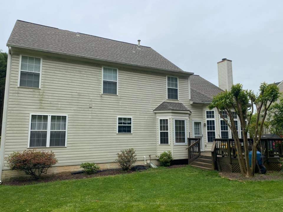 Back of a two-story beige house with a brown roof, windows, and a wooden deck on a grassy lawn.