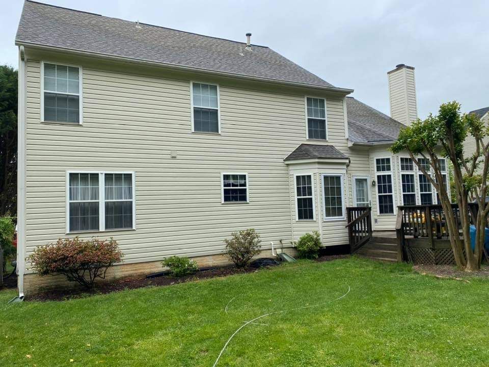 Back of a two-story beige house with a chimney, windows, and a wooden deck. Green grass and cloudy sky.