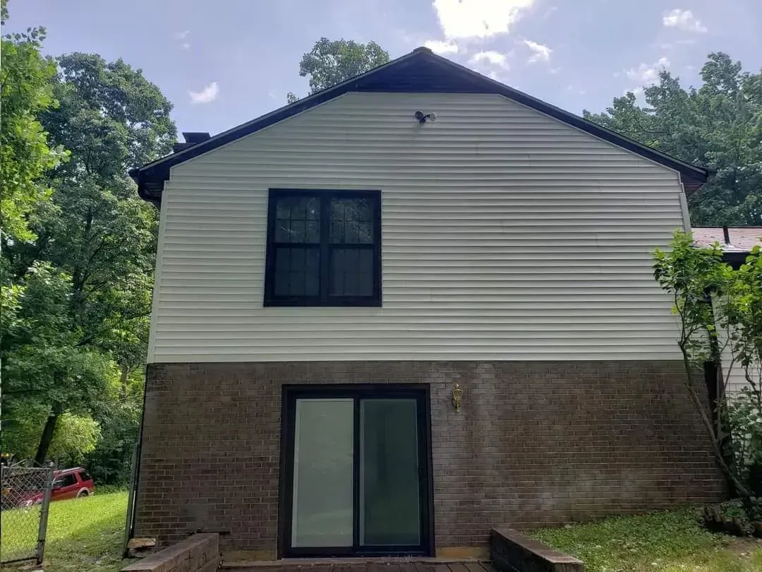 Back of a two-story house with white siding and a brown brick foundation, set in a wooded area.