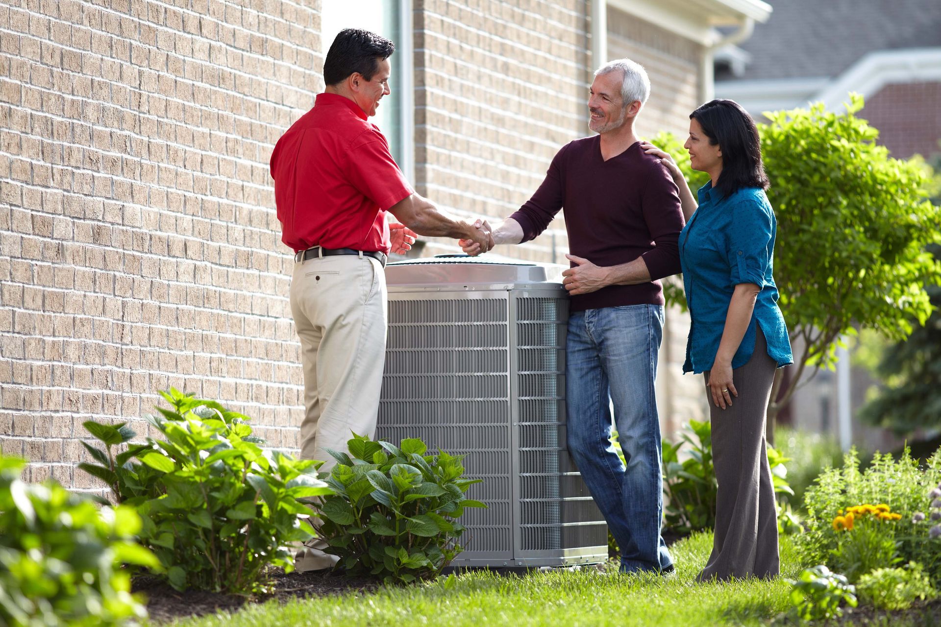 A man in a red shirt is shaking hands with a man and woman in front of an air conditioner.