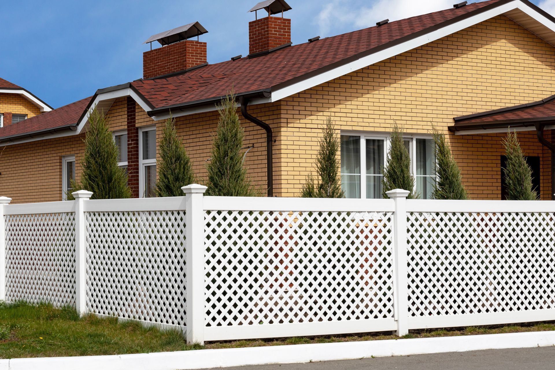 White lattice fence in front of a yellow house with a brown roof. Evergreen trees stand near windows.