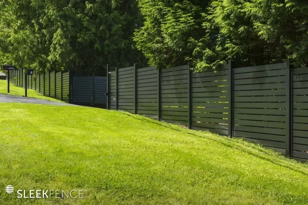 Dark gray horizontal slat fence along a grassy slope, with trees in the background.