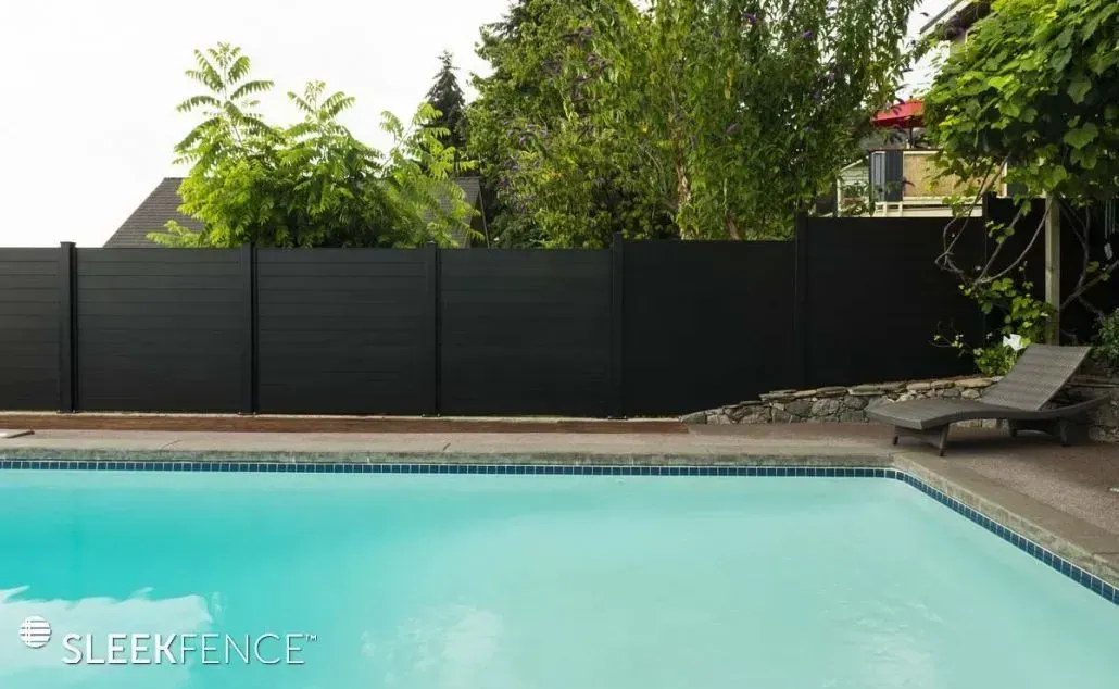 Black fence bordering a pool, with trees and a lounge chair in the background.
