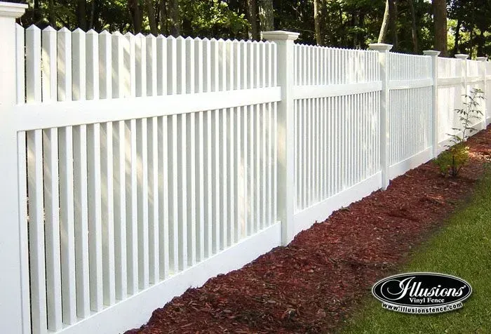 White picket fence with vertical slats bordering a red mulch bed and green grass.