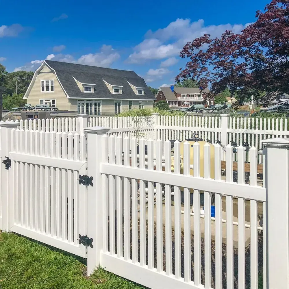 White picket fence surrounding a backyard with a house in the background on a sunny day.