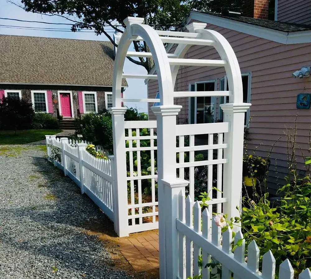 White arbor and picket fence leading to a pink building and a house with pink door.