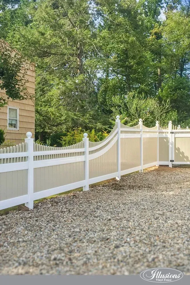 White vinyl fence along a gravel driveway, with trees in the background and a house on the left.