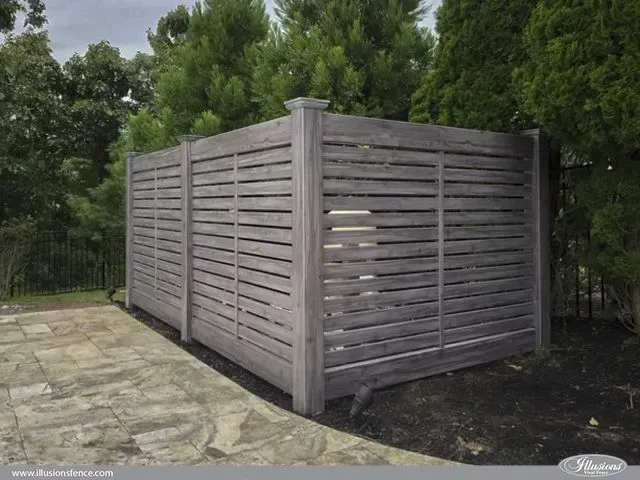 Wooden slatted fence in a backyard, surrounded by trees and a stone patio.