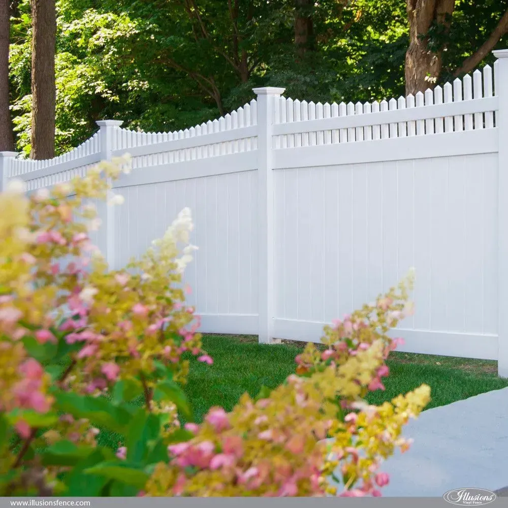 White picket fence in a yard, with yellow and pink flowers in foreground, trees in background.
