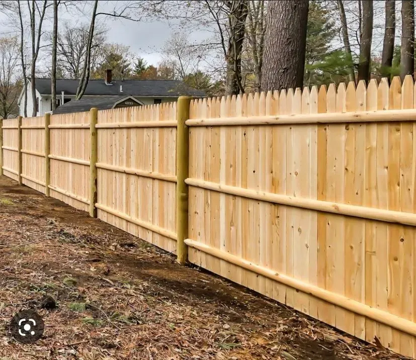 Wooden picket fence, cedar tone, enclosing a yard; posts are round, with a house in the background.