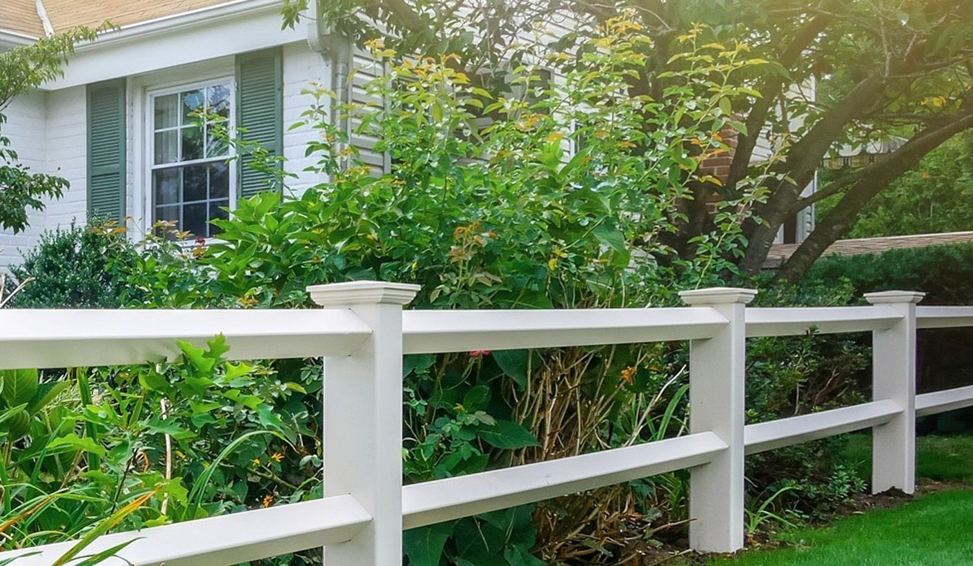 White fence in front of a house with green shutters, surrounded by lush greenery and a sunny backdrop.