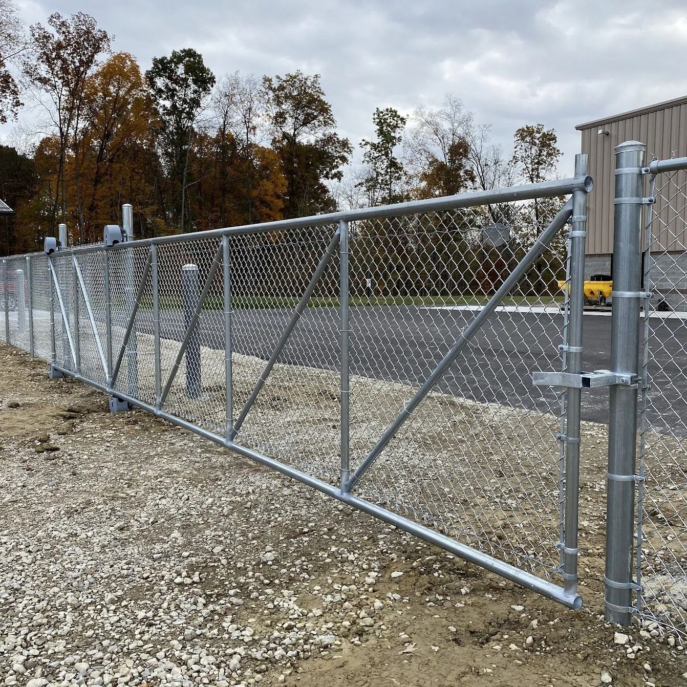 Chain-link fence with a large gate, set in gravel. Steel posts and a cloudy sky backdrop.