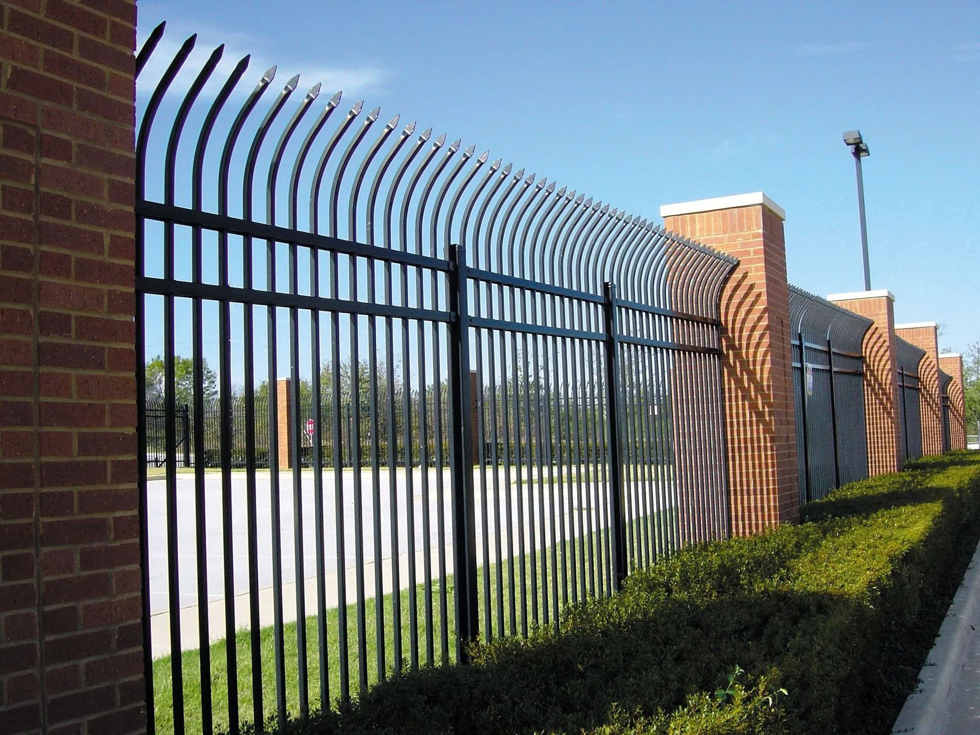 Black metal fence with curved top between brick columns, green bushes in front.